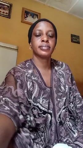 A woman sits indoors, wearing a black headscarf and a patterned robe over her clothes. The walls are yellow, and three framed pictures hang on the wall behind her, with a white refrigerator visible to the left. She looks directly at the camera and moves her hands slightly, appearing to be speaking. Loud music plays in the background.