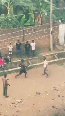 A daytime outdoor scene shows two men engaged in a physical altercation, likely boxing. The shirtless man throws punches at the man in a white shirt and shorts, who holds his left hand up in a guard and ducks slightly. The fight takes place on a dirt ground between two concrete channels. A small group of people, mostly young men, watch the fight from behind the concrete channel. An additional person wearing what looks like a military uniform stands on the left edge of the frame, also watching. A brick wall and a metal gate are visible in the background, along with greenery. Overhead wires cross the scene.