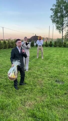 The video shows an outdoor setting with three people on a grassy field. One man, dressed in a dark suit, holds a microphone and a basket filled with food items. Another woman stands beside him wearing a light-colored pantsuit and holding a camera. In the background, there is a man wearing a yellow hat and light-colored pants, appearing to be joking or dancing. As the man in the suit begins to speak into the microphone, the camera zooms out and shifts focus. The man in the yellow hat is then seen walking towards another man dressed in brown pants and a green shirt who is holding a phone and recording. The background includes trees, houses, and a clear sky.