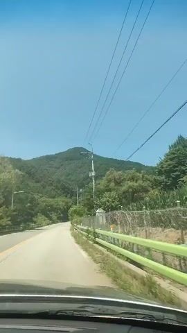 The video shows a first-person view from inside a car driving on a paved road. The road curves to the left and is surrounded by lush green trees and a mountain in the background. The sky is clear and blue. There are utility poles with wires running alongside the road. A green metal fence is visible on the right side of the road. The car is moving forward, and the scenery is passing by.