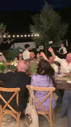 This video captures a lively outdoor evening celebration, likely a wedding reception, set in Montepulciano, Italy. The scene unfolds at dusk, with string lights illuminating the space and casting a warm glow on the festivities. Guests are seated at tables laden with floral arrangements, raising wine glasses in a toast.

The subjects are a group of people, appearing to be family and friends, engaged in a convivial atmosphere. They are dressed in smart casual attire and formal wear, suggesting a special occasion. Objects of note include wine glasses, dining tables, chairs, and decorative flowers. The background reveals a rustic stone building, lush trees, and hints of tents or marquees, indicating a rural or vineyard setting typical of the Tuscan countryside.

The primary activity is a toast, with multiple individuals holding their glasses aloft, their expressions conveying joy and camaraderie. The time of day is clearly evening, transitioning into night, and the weather appears pleasant, allowing for an outdoor gathering. The overall emotion conveyed is one of happiness and celebration, amplified by the clinking of glasses and the shared moment of merriment. No visible text is present in the video.