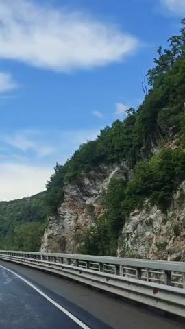 The camera records a car traveling on a wet asphalt road lined with white markings. Metal guardrails are visible on both sides of the road. A rocky cliff face covered in green vegetation rises on one side of the road. The sky is blue with scattered white clouds. As the car moves forward, the camera captures more of the landscape, revealing green hills and distant mountain peaks. Streetlights are seen lining the road. Another car is visible ahead, traveling in the same direction.