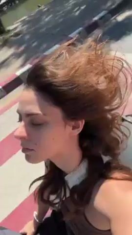 A young woman with curly reddish-brown hair rides on the back of a motorcycle. Her hair is blowing in the wind. The camera appears to be handheld by her. The background includes city buildings and trees, and people can be seen walking on the sidewalk.