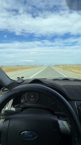 The video shows the interior of a car driving on a road. The dashboard and steering wheel are visible in the foreground. The road stretches out ahead, with a few other vehicles in the distance. The sky is blue with scattered clouds. The car appears to be moving at a steady speed. There are prayer beads hanging from the rearview mirror and a dashcam mounted on the dashboard. The radio or navigation screen shows an image of two people and some text. The overall impression is of a journey on a clear day.
