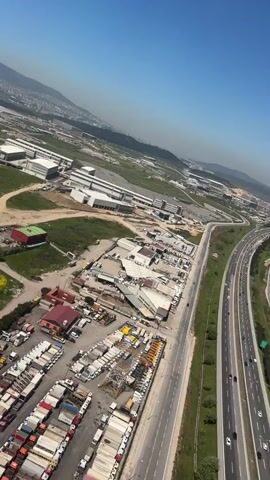 The video presents an aerial view of an industrial area on a sunny day. The foreground features rows of trailers and trucks parked in a lot, along with a building with a red roof. Adjacent to the lot, there is a grassy area with a patch of yellow flowers. Beyond this, a large industrial complex with multiple buildings is visible. In the distance, there are hills and what appears to be a city. To the right of the scene, a highway is visible, with traffic moving along it. The camera appears to be moving, slowly panning across the landscape. The sky is clear and blue.