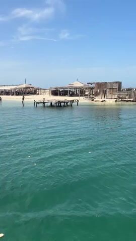 The video shows a wide shot of a tropical beach with clear turquoise water in the foreground. Several wooden structures with thatched roofs, likely restaurants or bars, are visible on the sandy shore. There are a few people walking on the beach and in the shallow water. A wooden pier extends into the water from the shore. The sky is blue with scattered clouds. The overall impression is a relaxed, sunny day at a beach resort.