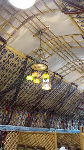 The video shows the interior of a yurt, a traditional nomadic dwelling. The yurt has a domed roof supported by wooden poles. The walls are lined with fabric featuring a blue and white pattern. Decorative tassels in various colors hang from the walls. A chandelier with multiple colored glass lamps hangs from the center of the roof. Wooden latticework divides the interior space, suggesting separate seating or dining areas. The overall impression is of a cozy, decorated interior with cultural influences.