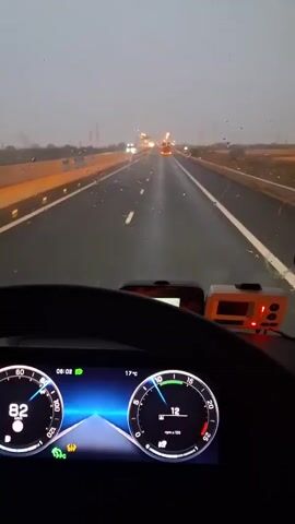 The video is shot from inside a truck cab, looking forward through the windshield. The wipers are active, indicating rain. The road ahead is wet and lined with white markings. The sky is overcast and appears to be dusk or dawn. The dashboard displays a speedometer reading of 12 and a tachometer. There are two electronic devices mounted on the dashboard. The camera pans slightly to the right, revealing the passenger side of the cab, including a photo frame with two pictures of children. The scenery outside consists of a highway with dry, grassy fields on either side.