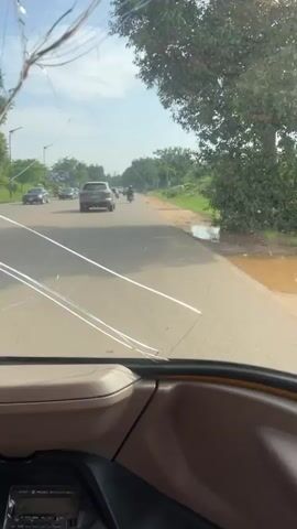 The video captures a view from inside a vehicle, looking out through the windshield and side window. The scene is a roadside with a paved road and a dirt shoulder. There is vegetation on the side of the road, including trees and grass. Several vehicles are visible on the road, including cars and motorcycles. The sky is cloudy. The windshield has visible cracks. The interior of the vehicle, including the dashboard and part of the steering wheel, is also visible.