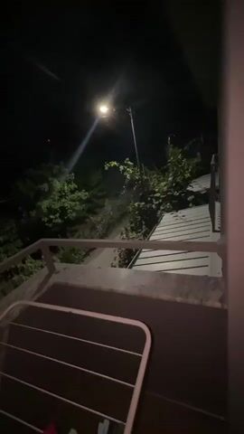 The video shows a view from a balcony or porch at night. A bright streetlamp illuminates the scene, casting long shadows. Lush green foliage is visible on a hillside behind a metal railing. A clothes drying rack is in the foreground. The camera pans slightly to the right, offering a wider view of the dark, wooded landscape. The streetlamp appears to be the only source of light, creating a stark contrast between the illuminated areas and the surrounding darkness.