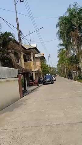 A person walks along a concrete road lined with palm trees and houses. A black SUV and a white car are parked on the side of the road. A red motorcycle is parked next to a house. Several people are visible in the distance, walking and standing. The sky is clear and blue.
