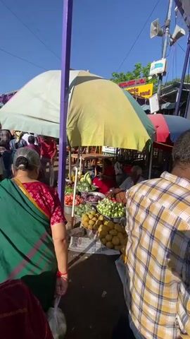 This is a vertical, wide-angle shot of a bustling outdoor market on a sunny day, likely in Chinnambedu, India, given the context. The scene is filled with people, predominantly women dressed in colorful sarees, browsing and shopping at various stalls. In the foreground, a woman in a green and red saree is facing away from the camera, holding a plastic bag.

The stalls are covered with large, brightly colored tarps, some white and some yellow, providing shade from the sun. These stalls display a vibrant array of fresh produce, including piles of mangoes, green gourds, leafy greens, tomatoes, and what appear to be onions or shallots. There are also baskets of fruit and vegetables. One stall has a man seated behind a display of produce, appearing to be a vendor.

The ground is dirt or pavement, with some newspapers scattered around the stalls. In the background, more stalls and people are visible, along with a tall utility pole with electrical wires. The sky is a clear, bright blue, indicating fair weather. The overall atmosphere appears lively and communal, typical of a local market where people gather to buy and sell goods. The time of day is clearly daytime, with strong sunlight casting shadows. There is some visible text on a sign in the background, but it is too blurry to read.
