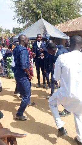 Several men are gathered outside, dancing in a circle on a dirt ground, as part of what appears to be a wedding celebration. Some of the men are wearing suits with bow ties, while others are in more casual attire, like a blue traditional outfit or a white robe.  A crowd of people, including women in colorful patterned dresses, are visible in the background. A blue and white canopy is providing shade, and a building with a corrugated metal roof is nearby. A car is also visible parked in the distance. The overall atmosphere seems festive and joyful.