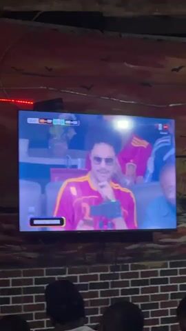The video shows a television screen displaying a soccer match, with an "HYDRATION BREAK" notification visible at the bottom. On the screen, a woman wearing large white sunglasses and a light blue jacket is seated next to a man wearing sunglasses and a gray suit. They are looking towards the field. The television is mounted on a brick wall, which is decorated with a mural of a ship and birds. In the foreground, the back of a person with dyed blonde hair is visible, along with the heads and shoulders of other people in the audience. Green LED lights are illuminated above the audience.
