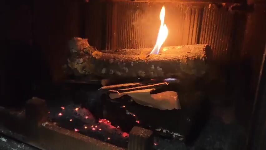 A still shot shows a fireplace with a burning log inside. A small flame rises from the log, and underneath it are pieces of paper or kindling, some of which are also burning. Embers glow at the bottom of the fireplace. The back of the fireplace is lined with a corrugated, metallic surface.