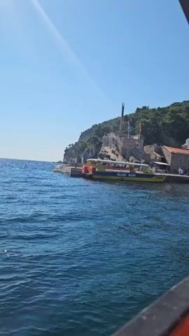 A yellow boat labeled "GLASS BOAT" is docked at a stone pier on a sunny day. The boat has people on it and appears to be a tourist vessel. Behind the pier, a rocky hill is covered with green vegetation and dotted with buildings, one of which is a stone structure with a red-tiled roof. A tall, slender monument or spire stands on the hill. The water in the foreground is deep blue and choppy. The sky is clear and bright blue. The camera angle suggests the footage is being filmed from another boat.