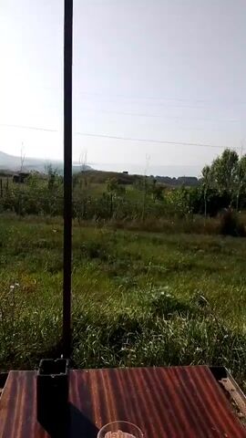 The video opens with a close-up of a wooden surface and a metal pole, with a green, grassy field and a bright sky in the background. As the camera pans right, it reveals more of the field, including some distant trees and hills. A metal structure with a corrugated roof supported by poles is visible in the foreground on the right side of the frame, suggesting an outdoor seating area or shelter. The overall scene depicts a rural or semi-rural environment on a clear, sunny day.