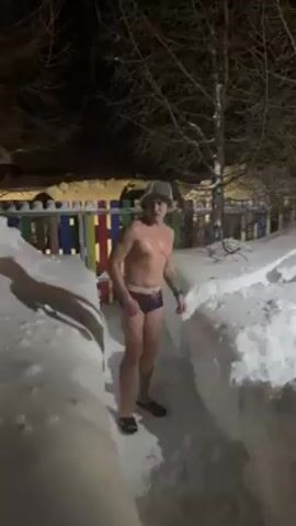 At night, a man wearing a bucket hat and underwear stands between two snow-covered paths, and then jumps into a pile of snow. The man is shirtless and also wears flip-flops. He disappears into the snow, makes a peace sign with his fingers, and then another person's arm reaches into the pile and pulls the man further into the snow. In the background, there is a fence made up of colorful vertical slats, trees, and an out-of-focus building.