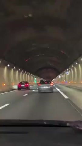 A car is driving through a tunnel. The tunnel is lit with evenly spaced lights along the walls. There are other cars in front of the camera's car, including a red car and a gray car. The road is marked with white lines. The tunnel's ceiling is arched.