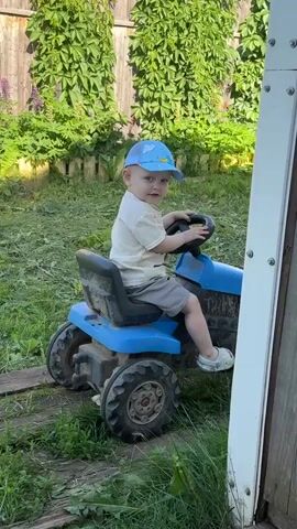 A young child, wearing a light-colored t-shirt, grey shorts, white sneakers, and a blue baseball cap with a cartoon design, sits on a blue toy tractor. The child is positioned behind the steering wheel, with their hands resting on it. The child looks directly at the camera and makes various facial expressions and hand gestures, including waving. The toy tractor is on a grassy area, with cut grass visible on the ground. In the background, there is a wooden fence covered in dense green vines, and some purple flowers. A portion of a white structure is visible on the right side of the frame. The scene appears to be outdoors on a sunny day.