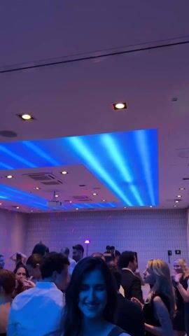 The video shows a group of people dancing in a room with blue and red lighting. They are generally well-dressed, and appear to be having a good time. Some are talking and laughing with each other, while others are focused on the music. There is an exit sign on the door.