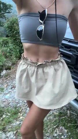 The video features a person, appearing to be a young woman, posing outdoors in Nậm Dịch Commune, Vietnam. She is dressed in a form-fitting gray strapless crop top and a high-waisted, ruffled beige mini skirt. Sunglasses are draped around her neck on a strap, and she wears a delicate necklace. Throughout the short clip, she makes subtle movements, swaying slightly and briefly adjusting her skirt with her right hand.

The setting is a natural, outdoor environment, captured during daylight hours with bright, natural light. The ground is a mix of dirt and gravel, with scattered green weeds and grass. A dark-colored vehicle, resembling a pickup truck or SUV, is partially visible behind her, showing its front grille and a headlight. A partial license plate displaying "179.83" is clearly seen on the vehicle. The background is filled with lush green trees and bushes, indicating a rural or mountainous landscape. No specific weather conditions beyond clear daylight are evident, and the person's expression is neutral, focusing on the pose.