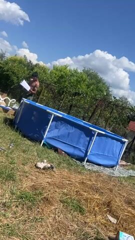 The video shows two young men assembling a blue above-ground swimming pool in a grassy yard on a sunny day. The pool is partially assembled, with a metal frame and the blue liner visible. One man is holding a set of instructions and looking at them, while the other is holding a piece of the pool liner. There are also cardboard boxes and pool frame pieces scattered around the area. In the background, there is a fence, trees, and a car.