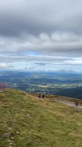 A vast, cloudy sky with patches of blue dominates the upper portion of the frame. Below the sky, a panoramic view of rolling hills and valleys unfolds. In the foreground, a grassy slope with dry, yellowed grass slopes down to a winding dirt path. Several people are visible on the path: two individuals stand closer to the camera, while further along, a pair walks away from the viewer, and another single person walks toward the camera. The landscape is dotted with trees and scattered buildings, indicating a rural or semi-rural area. The overall scene depicts a scenic, outdoor environment, likely a mountain or high vantage point, under a dynamic sky.
