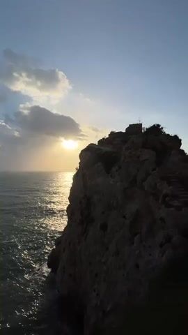 This video captures a scenic coastal view in Terrasini, Italy, during what appears to be sunset or sunrise, given the warm, golden light illuminating the scene and the soft glow in the sky. The primary subject is a dramatic cliff face that juts out into the sparkling, turquoise sea. The rocky terrain is rugged and textured, with a weathered appearance.

A prominent feature of the cliff is a set of stone stairs, winding their way up towards a small structure or ruins at the very top. These stairs suggest a path or access point to the summit, hinting at a historical or religious site, possibly a small chapel or lookout tower, indicated by a cross-like silhouette at the peak. The surrounding vegetation is sparse, with hardy, coastal plants clinging to the rocks and the edges of the path.

The sea is dynamic, with gentle waves lapping against the base of the cliffs, reflecting the soft light of the sun. The sky is a clear blue, with a few wispy clouds adding depth and texture to the upper portion of the frame. The overall atmosphere is tranquil and serene, evoking a sense of peace and natural beauty. The video pans slightly, offering different perspectives of the cliff, the sea, and the path leading towards it. There is no visible text in the video.