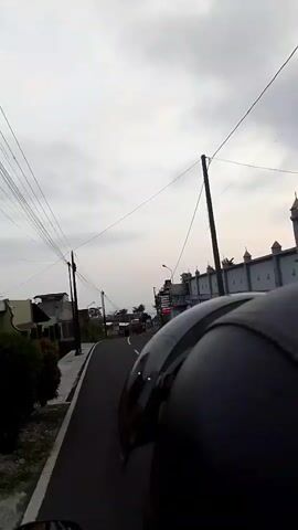 The video shows a first-person perspective of someone riding a motorcycle on a paved road. The road is curved and slopes slightly downhill. On the left side of the road, there is a sidewalk, some vegetation, and houses. On the right side of the road, there are more houses, some with decorative architectural elements, and a billboard advertising "SiMBa Top". Overhead, there are many power lines strung between poles. The sky is overcast with grey clouds. The camera is focused on the road ahead and the rider's helmet, which is black. The rider is wearing a helmet.