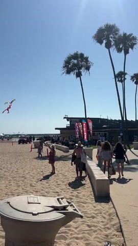 A kite is visible in the sky above a sandy beach. Several people are scattered across the sand, some sitting under umbrellas and others walking. A volleyball game is in progress near the center of the frame. Palm trees and a building with banners are visible in the background. The sky is clear and blue.
