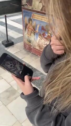 A person wearing a gray hooded sweater stands in a room looking at a wall with several paintings of biblical figures and creatures on it, taking pictures with a cell phone. The paintings appear to be quite old and have some signs of wear. The phone is a modern smartphone with a gold-colored case. The camera then pans up to show more detailed paintings on the walls and ceiling of the room, depicting various religious scenes with multiple figures. The ceiling is domed, and the paintings appear to be part of a larger, intricate design covering the entire surface.