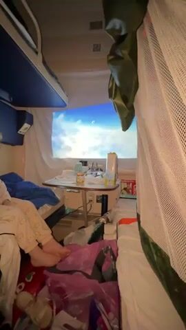 The video shows the interior of a train compartment with multiple people. The compartment appears to be set up for sleeping, with bunk beds visible. On one wall, a projection screen displays a cartoon image, possibly for entertainment. There is a small table with various items on it, including jars and a box. In the foreground, a person's legs are visible, and their feet are bare. There are blankets and pillows on the beds. Some luggage or bags are present on the floor. The lighting is dim, suggesting it may be nighttime or the lights are turned down. The overall scene depicts a family or group traveling by train, with efforts made to entertain a child or children.