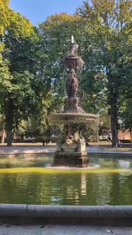 The video shows a classic tiered fountain in a park. The fountain is made of stone and features statues of figures. Water cascades down from the top tier into a lower basin, and then into a larger pool surrounding the fountain. The pool's water is a murky green color. Lush green trees with full canopies surround the fountain, and the sky above is a clear blue. The sun is shining, casting shadows and highlighting the water's movement. There are a few people in the background, walking or standing, but they are out of focus and their actions are indistinct. The overall scene depicts a serene park setting with a prominent water feature.