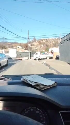 The camera faces forward from inside a vehicle, positioned at a stop on an asphalt road. In the foreground is the dashboard, covered with a pile of papers. Through the windshield, a white flatbed truck is parked to the right. Two people are visible in the bed of the truck. Behind the truck, the landscape includes houses and vegetation on a hillside. Many of the trees appear to have been burned. A utility pole is on the left side of the street. The sky is clear and blue.
