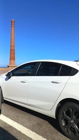 The video shows a brick chimney standing near the ocean. There's a paved area surrounding the chimney, with benches where people are sitting. A bike path runs alongside the paved area, and a cyclist passes by. The sky is clear and blue. A side mirror is visible in the corner of the screen.