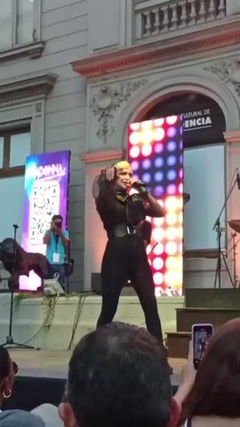 A female performer in a black jumpsuit with sheer sleeves sings into a microphone and dances on an outdoor stage with a band behind her. The backdrop features a bright light display and banners with the word “MADONNA” and QR codes. The audience films and watches, some clapping. A cameraman stands to the side. The stage is set against an old, light-colored building with decorative architectural details.