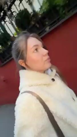 A young woman with fair skin and light brown hair in a low ponytail is walking outside. She is wearing a white, fluffy sherpa jacket with a brown strap of a bag slung over her shoulder. The background consists of a red painted wall with iron fencing and foliage, suggesting a residential or urban environment. The camera appears to be hand-held and follows her as she walks and looks around. The lighting indicates it is daytime.