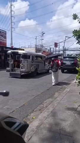 The video opens with a view of a street scene, possibly in the Philippines given the presence of a jeepney, a common form of public transportation there. The shot is taken from the perspective of a motorcycle, with part of the handlebars and mirror visible in the foreground. The street is paved and appears to be in a commercial area, with various shops lining the side of the road.

In the distance, a gray jeepney is driving away from the camera. It has passengers visible inside and a metal frame extending from the back. A person with a white shirt and backpack is walking away from the jeepney towards the right side of the frame.

To the left of the street, there are several storefronts. One sign reads "MINUTE BURGER." Another sign, partially obscured, appears to advertise mobile phone services or products, with images of people and text that includes "1Gbps." Further down the row of shops, a prominent sign displays "DUNKIN'." People are seen entering and exiting these establishments.

The sky is blue with scattered white clouds, suggesting daylight. Power lines and utility poles are visible overhead. The overall atmosphere appears to be that of a bustling urban or suburban street on a sunny day. The camera pans slightly throughout the video, offering different angles of the street, shops, and moving vehicles.