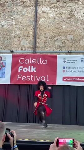 This video captures a lively folk dance performance at the Calella Folk Festival. The central subject is a dancer in traditional attire, featuring a red tunic with intricate white embroidery, dark leggings, and tall black boots. Their movements are dynamic, including jumps and spins, suggesting a vigorous and energetic dance style. The dancer wears a dark, curly wig and a hat, adding to the traditional costume.

The performance takes place on a stage in front of a large red banner that reads "Calella Folk Festival." The banner also displays the logo of the Ajuntament de Calella and social media handles for "Folklore_Fes" and a website address "www.folklore...". To the left of the banner, a small image of a person in traditional costume is visible. Behind the stage, a rough stone wall forms the backdrop, suggesting an outdoor setting possibly in a historic area. The stage is covered with black curtains, and there are lights casting red and green hues on the stage floor.

In the foreground, two audience members are actively filming the performance with a DSLR camera and a smartphone. This indicates the event is being documented, likely by festival attendees or media. The lighting suggests it might be daytime or early evening, as the natural light from the stone wall is visible, but the stage lights are also prominent. The overall atmosphere appears festive and engaging, with the dancer's animated performance drawing the attention of the audience.