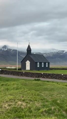 A vertical shot captures the iconic black church at Búðir, Iceland, under an overcast sky. The church, Búðakirkja, stands prominently in the midground, characterized by its dark wood siding, a crisp white double door, and white-framed windows. A steeple with a cross and a weathervane crowns the structure. To the left of the church, a tall white flagpole stands vertically.

In the foreground, vibrant green grass covers the landscape, with a path or dirt road running horizontally in front of a low, dark stone wall that separates the grassy area from the church grounds. Beyond the church, the terrain is also green, leading up to a dramatic backdrop of rugged mountains. These mountains are partially shrouded in clouds, and some peaks are visible with patches of snow or ice. The sky above is entirely covered with grey, thick clouds, creating a diffused and somewhat solemn light. There is no visible human activity or text in the image. The scene conveys a sense of serene isolation and raw natural beauty characteristic of Iceland's landscapes.