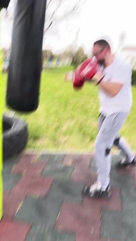 Here is a description of the video. 
The video shows a man boxing a punching bag in an outdoor setting. The man is wearing boxing gloves, a white t-shirt, grey sweatpants, and glasses. He is standing on a red and grey tiled surface. The punching bag is black and cylindrical. In the background, there is a green lawn, a bare tree, and a white building with a bell tower. The sky is overcast.