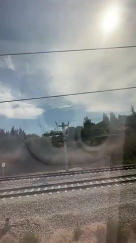 The video shows a view from inside a moving train, looking out the window. The sun is bright and partially obscured by clouds in a blue sky. Outside, there are train tracks and gravel.  A low, dry, grassy hill with sparse trees runs along the tracks. There's a low concrete retaining wall alongside the tracks. Overhead, there are electrical wires and support structures for the train's power system. Streaks and smudges are visible on the window.