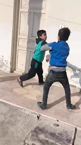 Two young boys are playing in an outdoor, paved area. The boy on the left wears a blue long-sleeved shirt and dark jeans, with his underwear showing. The boy on the right wears a green-and-black sweatshirt and black pants, with brown sandals. They playfully fight near a white wall.  The boy in green kicks at the other boy, then they briefly grapple. A white door and a metal-framed window are visible along the wall. The sun casts their shadows on the wall.