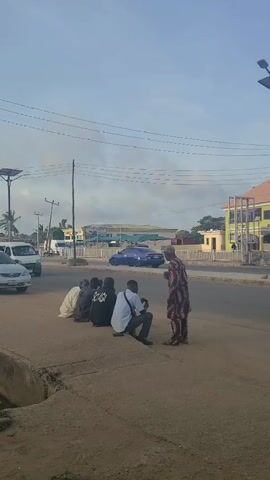 The video shows a group of four men sitting on a sidewalk area next to a road while an older man wearing a patterned shirt and pants stands in front of them and appears to be talking to them. Behind the group, there is a road with moving traffic, including cars, vans, and auto rickshaws. In the background, there are buildings, including a multi-story building with a distinctive architectural design. Power lines are visible overhead against a somewhat cloudy sky. The overall setting appears to be a commercial or residential area in a developing country.
