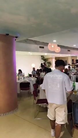 The video shows a wide shot of a dimly lit event hall with round tables covered in white tablecloths. Several groups of people are seated at the tables, engaged in conversation or eating. Two ornate chandeliers hang from the ceiling. A man in a light-colored t-shirt and shorts walks away from the camera towards the right side of the frame, while another man in a pink shirt with a backpack walks towards the camera on the right. In the background, several individuals are gathered around a stage area where a woman is speaking. The overall atmosphere appears to be a formal gathering or celebration.