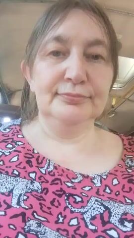 A close-up, waist-up video captures a middle-aged woman with light brown hair pulled back in a ponytail. She is wearing a pink shirt with a black and white leopard print. Her head is centered in the frame, and she is looking directly at the camera with a neutral expression. The background is slightly blurred, showing what appears to be the interior of a vehicle, possibly a bus, with a yellow pole visible above her head. The lighting suggests it is daytime, with some natural light entering the space. There are no other people or significant objects in the frame. The video is short, approximately four seconds long, and shows no movement or actions by the woman.