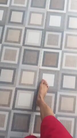 A person's bare feet and legs are visible on a tiled floor. The tiles are square and have a pattern of concentric squares in various shades of beige, gray, and white. The person is wearing red shorts. The camera then pans up to reveal a black metal gate with vertical slats. Behind the gate, there are green plants and trees. Sunlight casts shadows on the tiled floor. The video ends with a view of the gate and the surrounding foliage.
