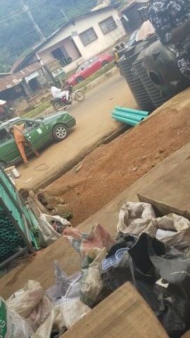 The media file captures a daytime street scene in Ado Ekiti, Nigeria.

In the foreground, a person wearing orange overalls or a garment is actively working on a green sedan. The individual is bent over, seemingly attending to the vehicle's underside or side, with its rear wheel prominent. The car is parked on the unpaved, reddish-brown shoulder of what appears to be a local road. Light blue PVC pipes are scattered on the ground near the vehicle, suggesting ongoing construction or material storage.

Further down the road, a red car and a white van are parked. On the left side of the street, several multi-story buildings, possibly residential or mixed-use, are visible, featuring multiple windows. At the upper left corner of the frame, two large, black water storage tanks are stacked. Overhead power lines crisscross the background, indicating local infrastructure.

To the right of the green car, a dirt embankment rises, cluttered with various items. These include large, white woven sacks, some appearing to contain waste or other materials, with fragmented and largely unreadable text visible on one. More light blue PVC pipes are also seen here, alongside wooden planks and green corrugated metal sheeting, possibly part of a makeshift structure or a roof. The ground in this area also shows signs of debris.

The lighting suggests it is daytime, and the overall atmosphere is one of daily activity in a semi-urban or urban environment. No specific emotions are discernible from the individuals.