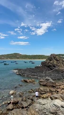 This video shows a scenic coastal view on a sunny day. The camera is positioned above a rocky shoreline, looking out towards the ocean. In the foreground, two people are exploring the rocks near the water's edge.

The water is a clear turquoise, with sunlight glinting off its surface. Numerous small fishing boats are scattered across the bay, appearing to be anchored or gently drifting. The distant shoreline is lined with lush green hills, meeting the bright blue sky dotted with fluffy white clouds.

To the right, a distinctive rock formation juts out into the sea, featuring layered, dark, columnar basalt. A few more people can be seen gathered on this rocky outcrop, observing the view. The overall impression is one of a peaceful, natural landscape, likely a popular spot for recreation and enjoying the coastal environment. The lighting suggests it is daytime, likely late morning or early afternoon given the bright sun. The activity depicted is leisurely exploration and observation of the beautiful scenery.